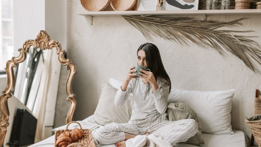 Woman in pajamas enjoys coffee and pastries in a stylish and cozy bedroom setting.